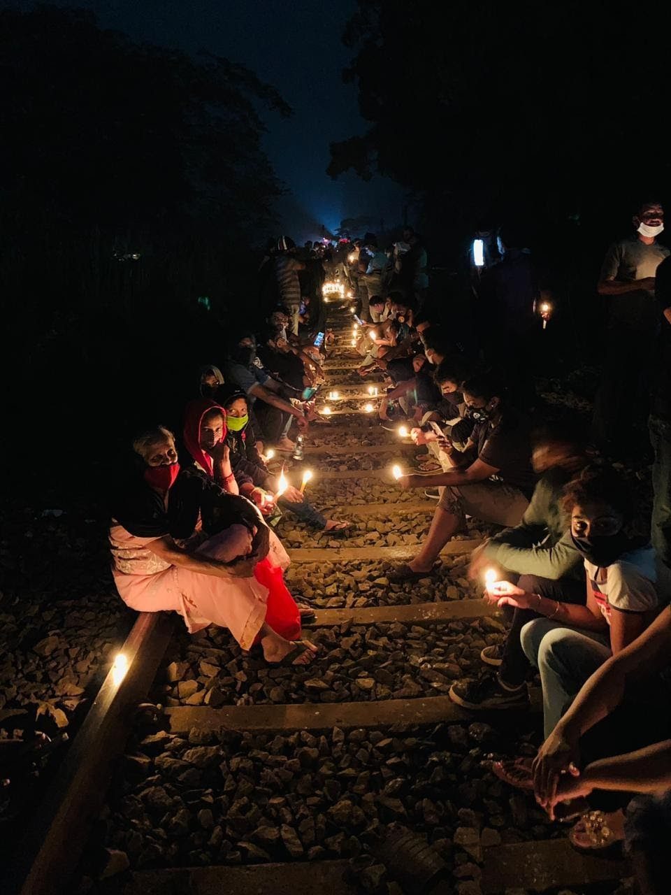 People sitting on both sides of the railway track at night, holding candles